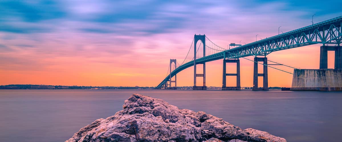 A suspension bridge over calm water at sunset with rocky foreground.
