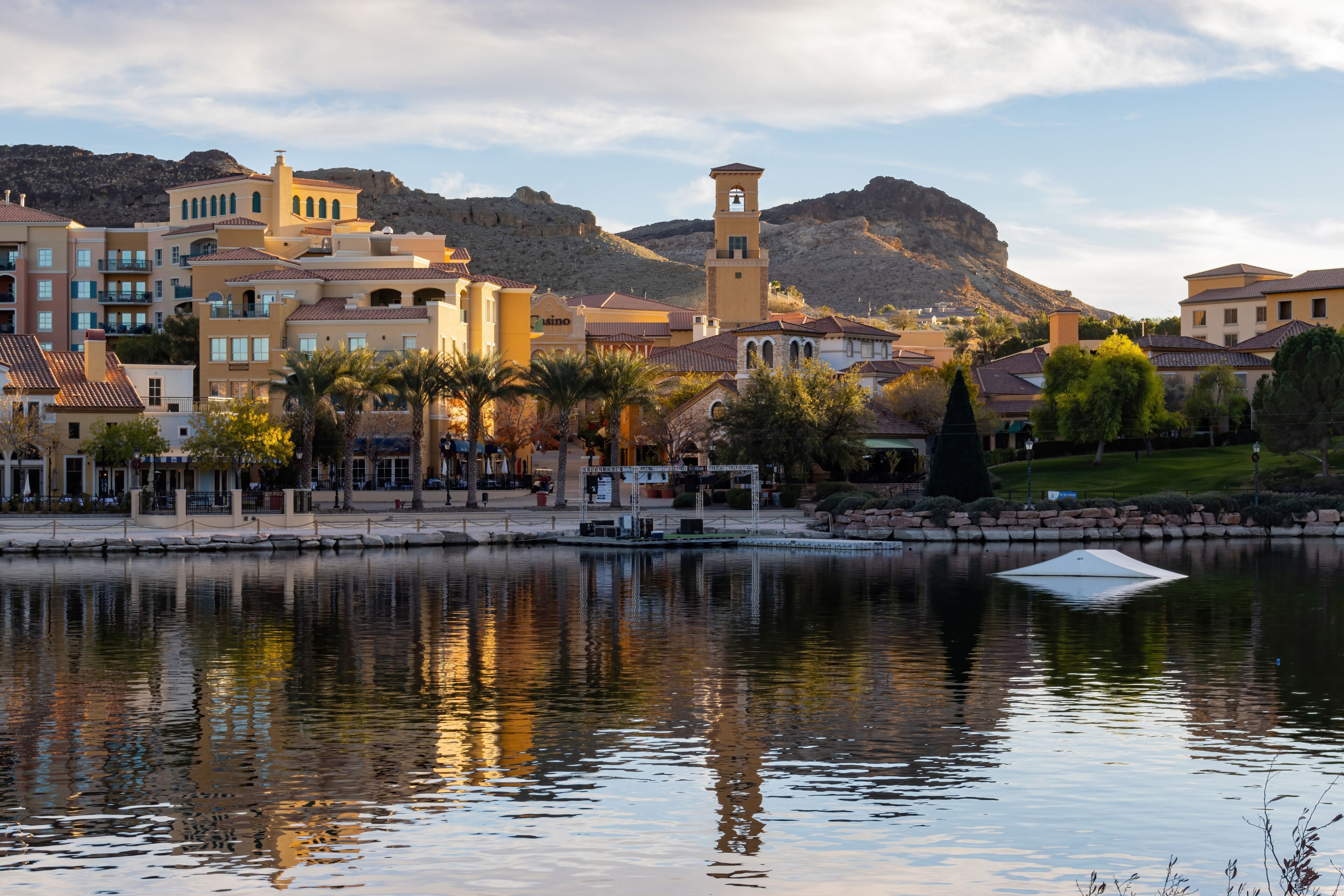 A lakeside town with Mediterranean architecture, a prominent tower, and mountains in the background.