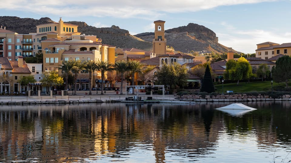 A lakeside town with Mediterranean architecture, a prominent tower, and mountains in the background.
