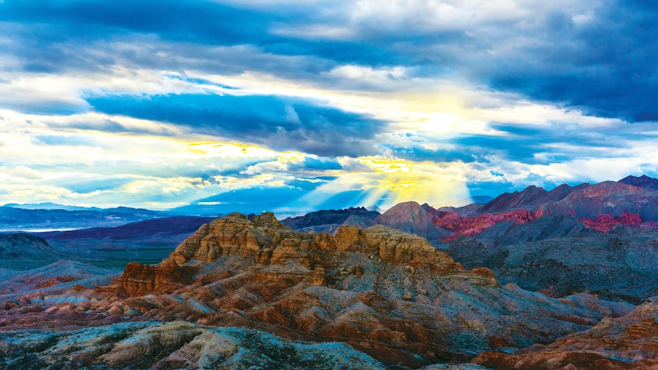 A rugged mountain landscape with a prominent rock formation in the foreground and a range of mountains in the background under a dramatic sky.