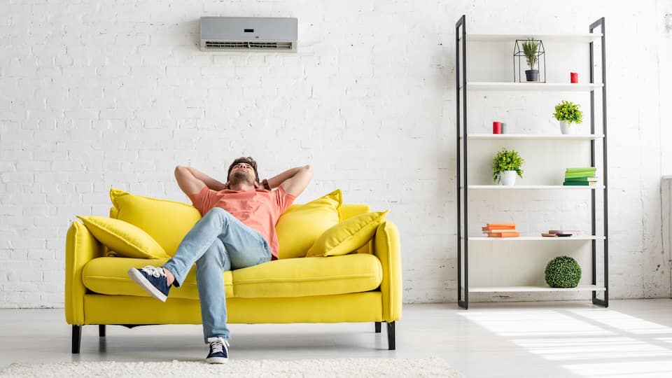 A person reclining on a yellow sofa in a room with a white brick wall, a white floor, and a white bookshelf with books and plants.