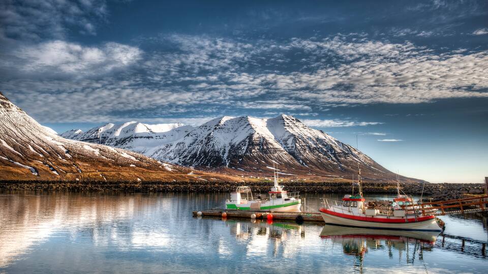 A calm fjord with two boats docked, a snow-capped mountain in the background, and a clear sky.