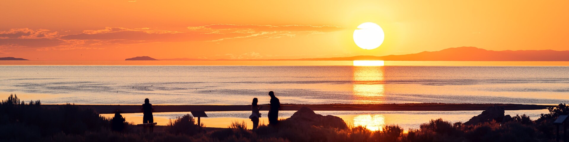 A sunset over a calm body of water with silhouettes of people and a distant shoreline.