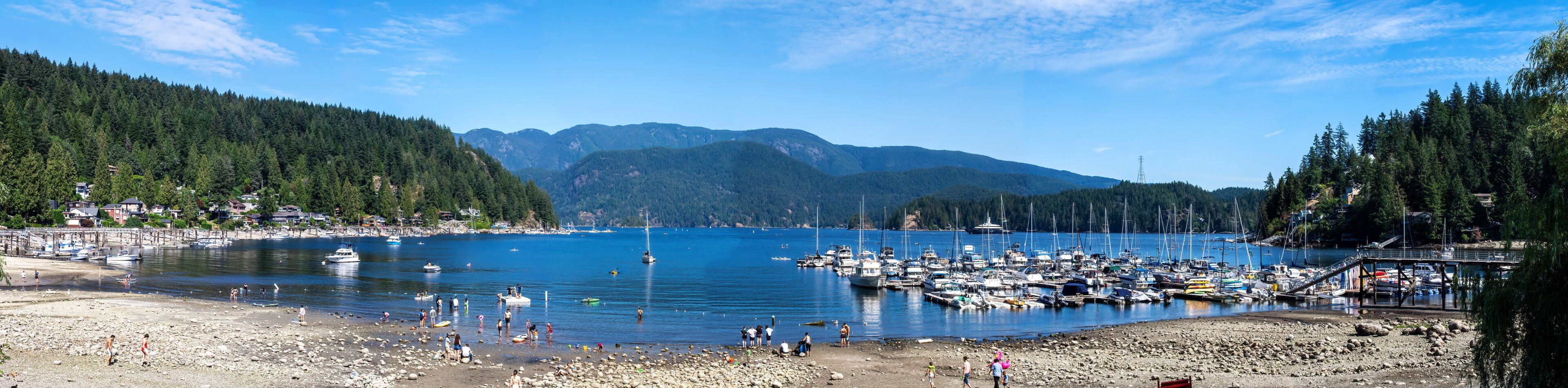 Una playa rocosa con gente, un puerto deportivo con barcos y montañas boscosas al fondo.