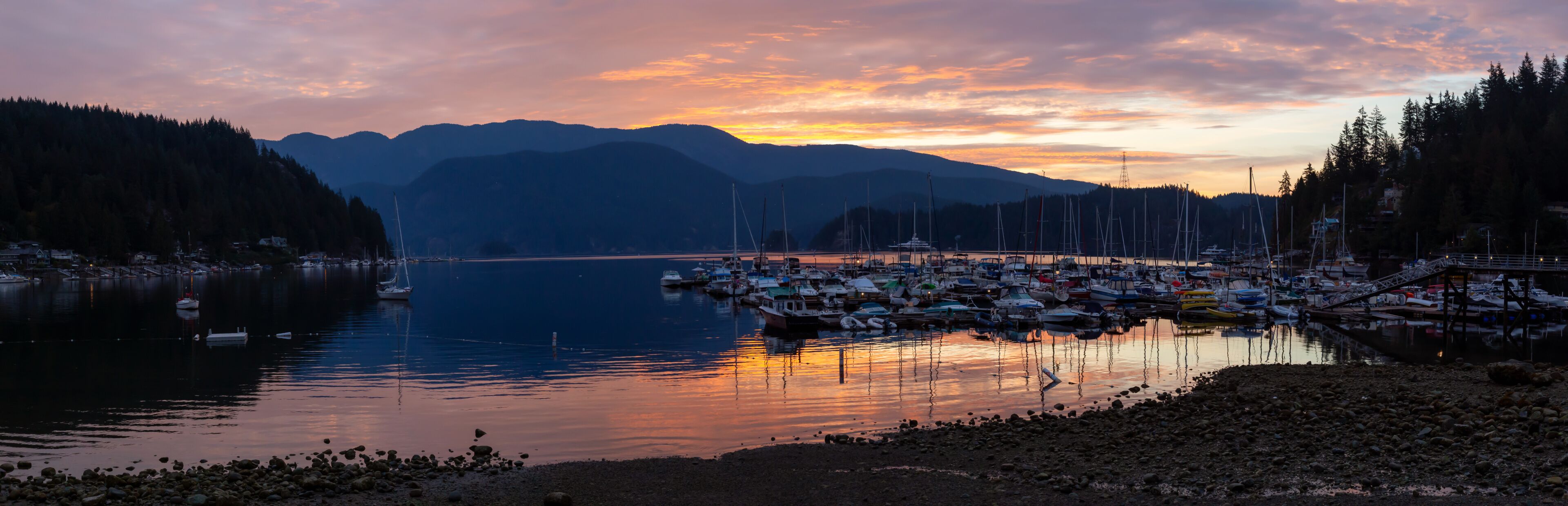 A marina with boats docked, surrounded by mountains and a sunset sky.