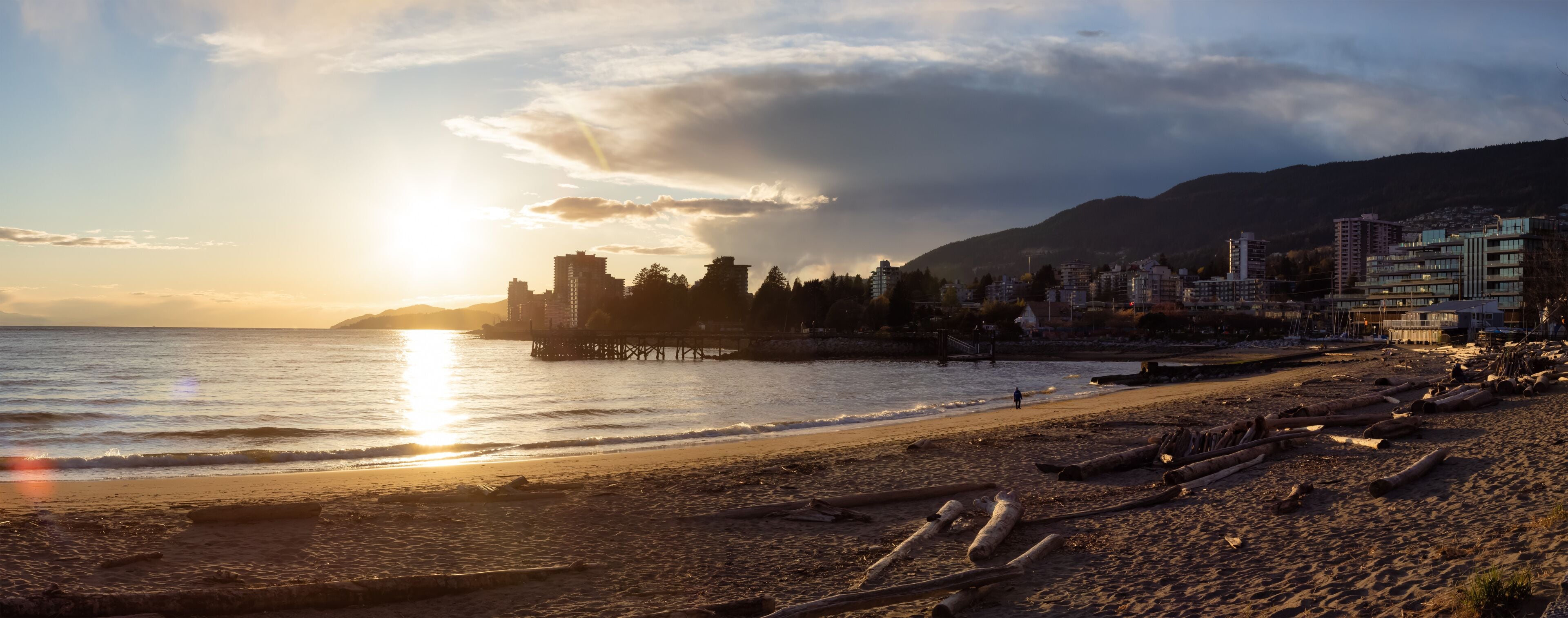 Una playa con un muelle, edificios y montañas al fondo.