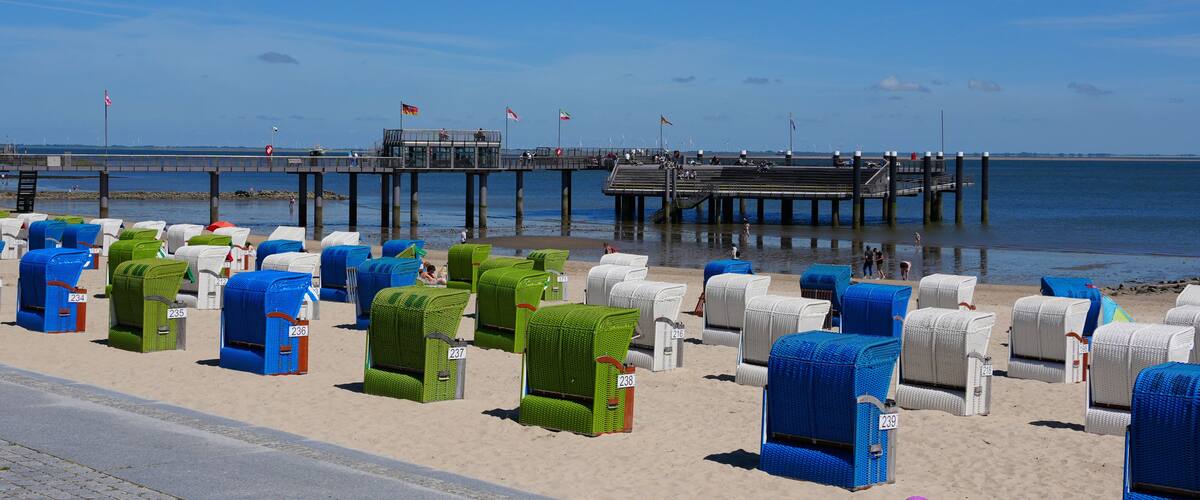 A lively coastal scene in Wyk auf Föhr, Germany. Tourists and locals relaxing on the sandy beach and promenade next to traditional strandkorb chairs and the famous Mittelbrücke pier at the North Sea.