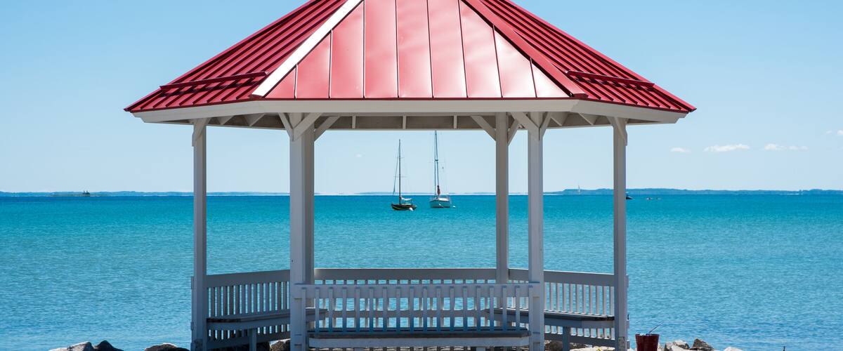 A white gazebo with a red roof overlooking a calm sea with sailboats.