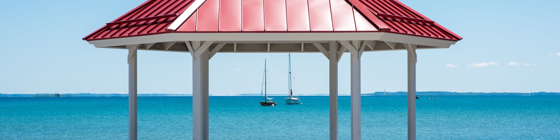 A white gazebo with a red roof overlooking a calm sea with sailboats.