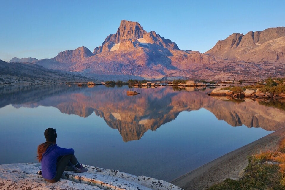A person sitting by a calm lake with mountains in the background.