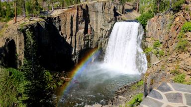 Una cascada con un arcoíris, rodeada de árboles y terreno rocoso.