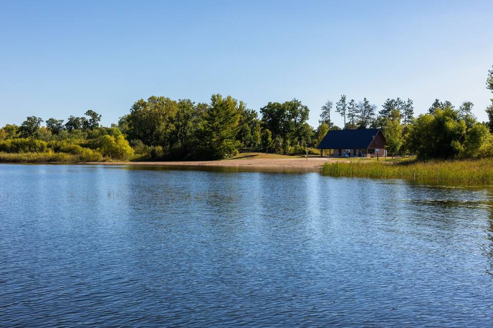 A calm lake with a wooden dock, surrounded by dense trees and a small building with a red roof.