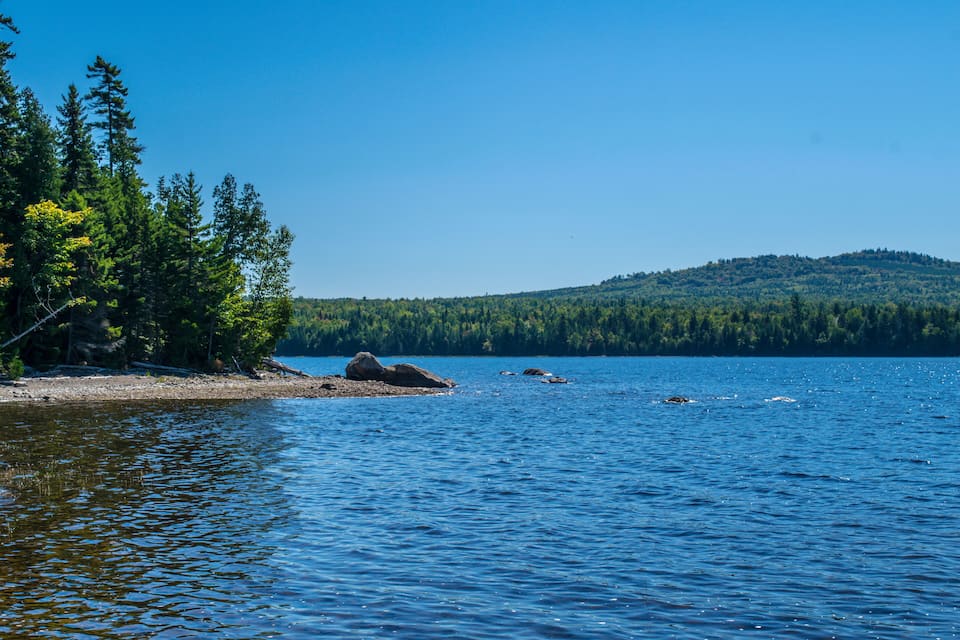 Un lac paisible aux eaux d’un bleu limpide, entouré d’une forêt verdoyante et dense, et dominé par une colline lointaine.