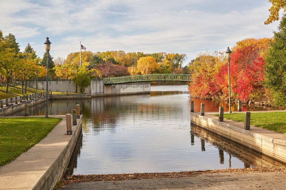 A calm river with a green bridge, autumn trees, and a flag on a pole.
