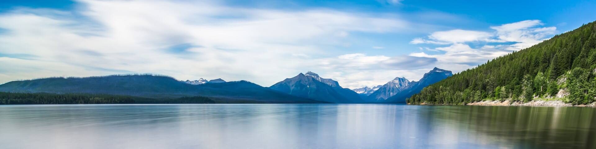 A clear lake with pebbles at the bottom, surrounded by mountains and trees.