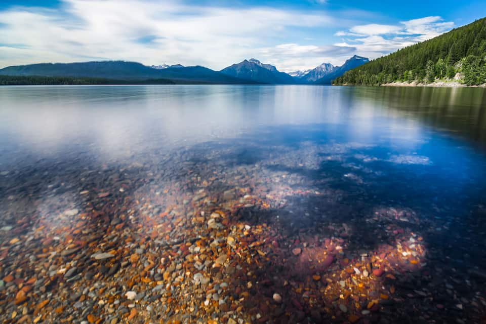 A clear lake with pebbles at the bottom, surrounded by mountains and trees.