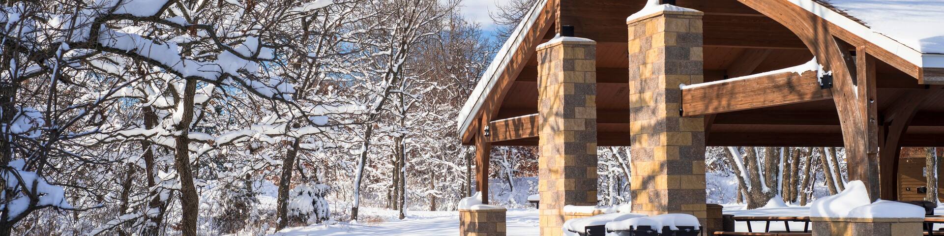 Un pavillon enneigé, doté de piliers en pierre et d’un toit en bois, entouré d’arbres sans feuilles et surplombant un ciel bleu clair.