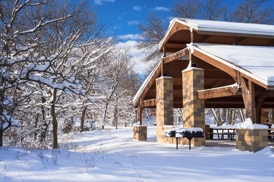 Een met sneeuw bedekte pagode met stenen zuilen en een houten dak, omringd door kale bomen en een helderblauwe lucht.