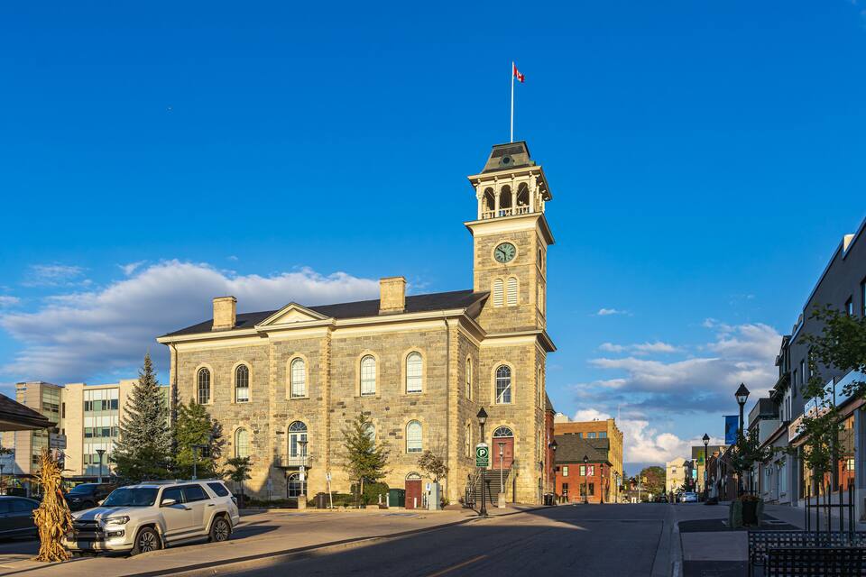 A historic stone building with a clock tower and a flag, surrounded by modern buildings and parked cars.