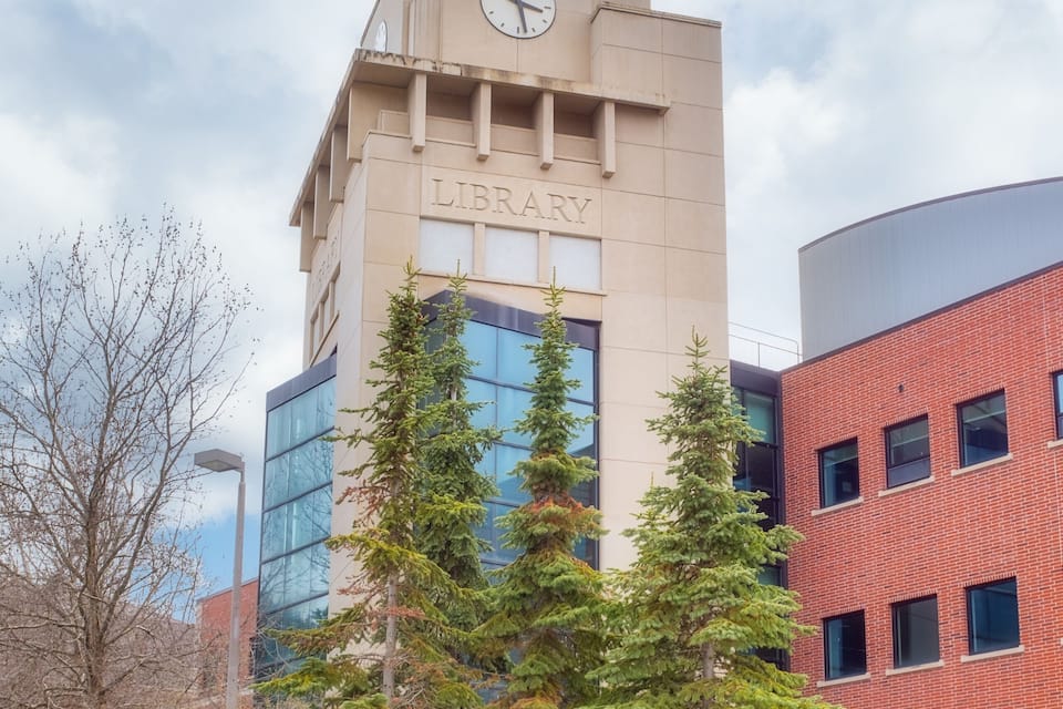A library with a clock tower, surrounded by evergreen trees and a circular flower bed.