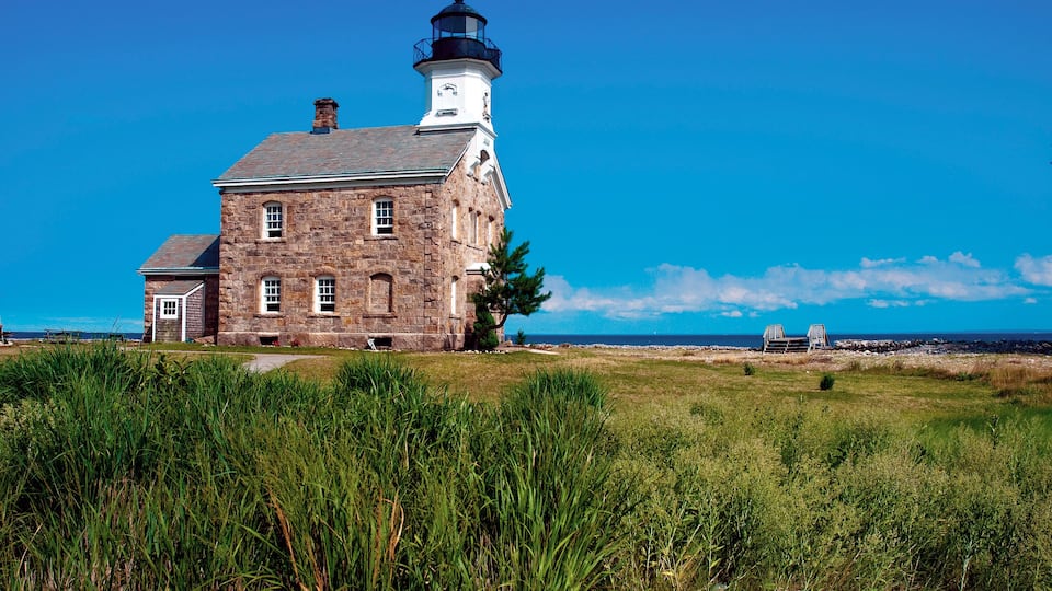 A stone lighthouse with a white tower and black lantern room, situated on a grassy cliff by the ocean.