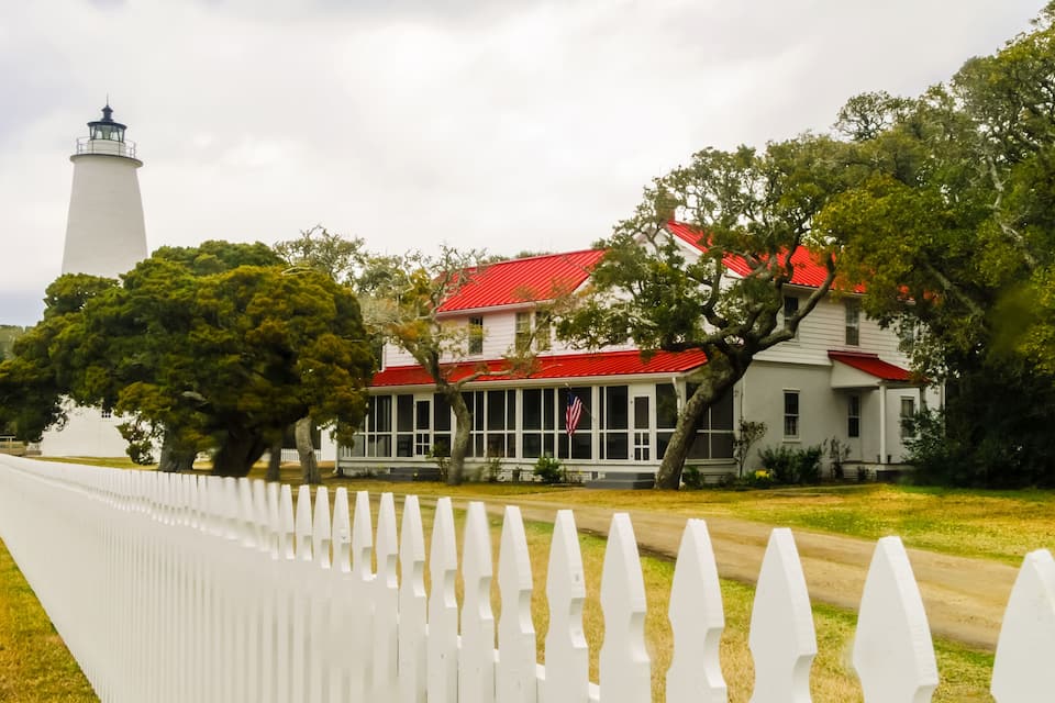 A white lighthouse, a red-roofed house, and a white picket fence.