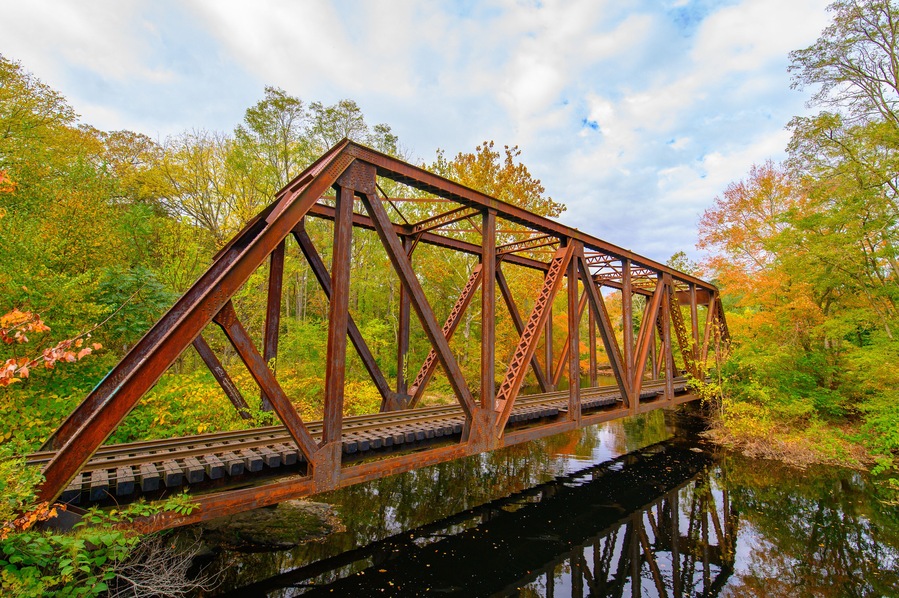 A rusted metal bridge over a calm river surrounded by autumn foliage.