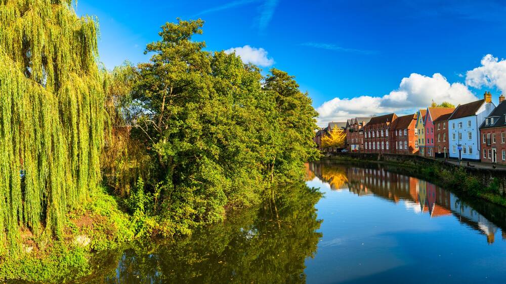 A calm river reflecting colorful buildings, a weeping willow, and a church spire.