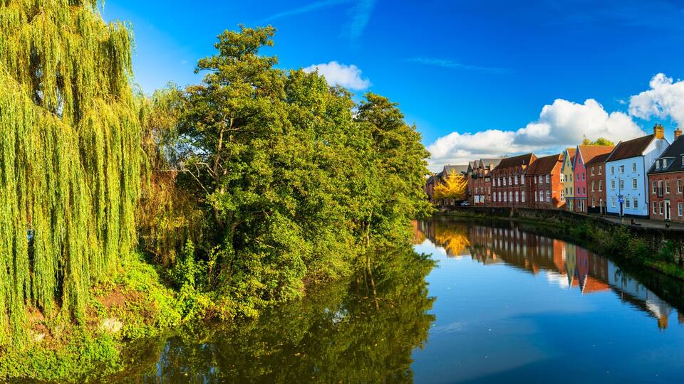 A calm river reflecting colorful buildings, a weeping willow, and a church spire.