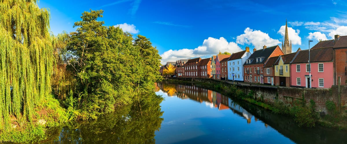 A calm river reflecting colorful buildings, a weeping willow, and a church spire.