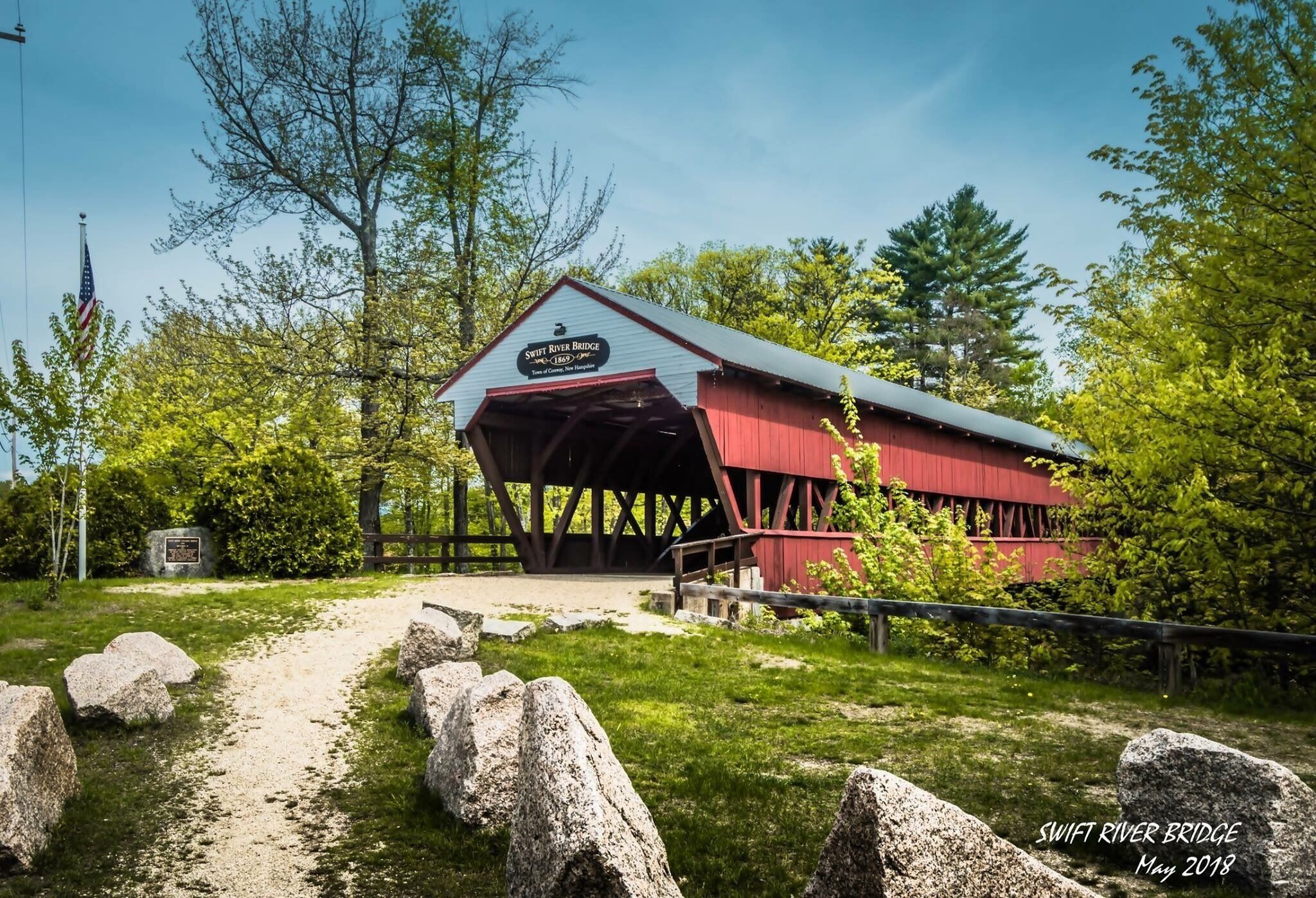 A red covered bridge with a sign reading "Swift River Bridge" in a wooded area.