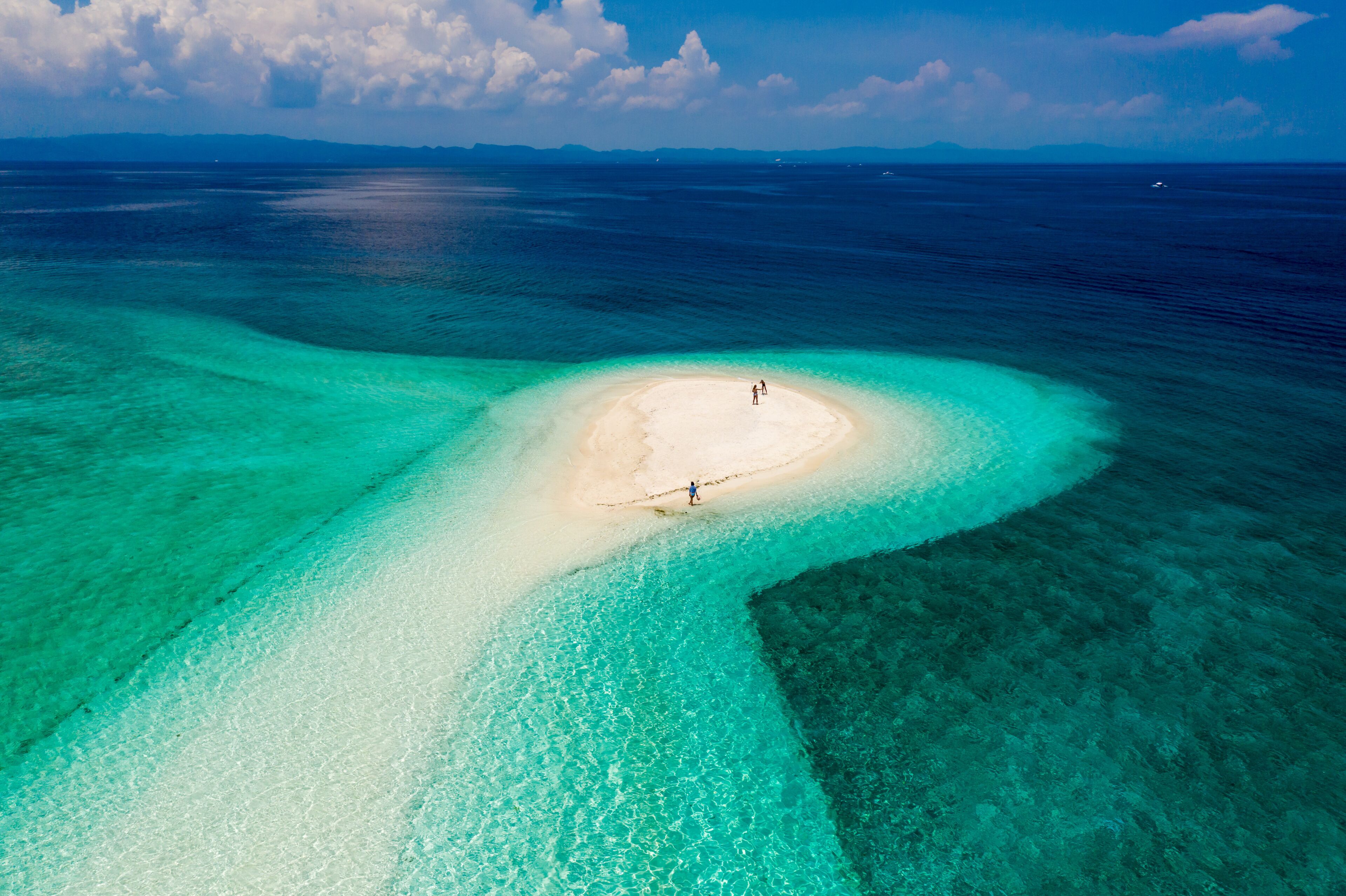 A clear aerial view of a turquoise lagoon with a sandy islet and two people standing on it.