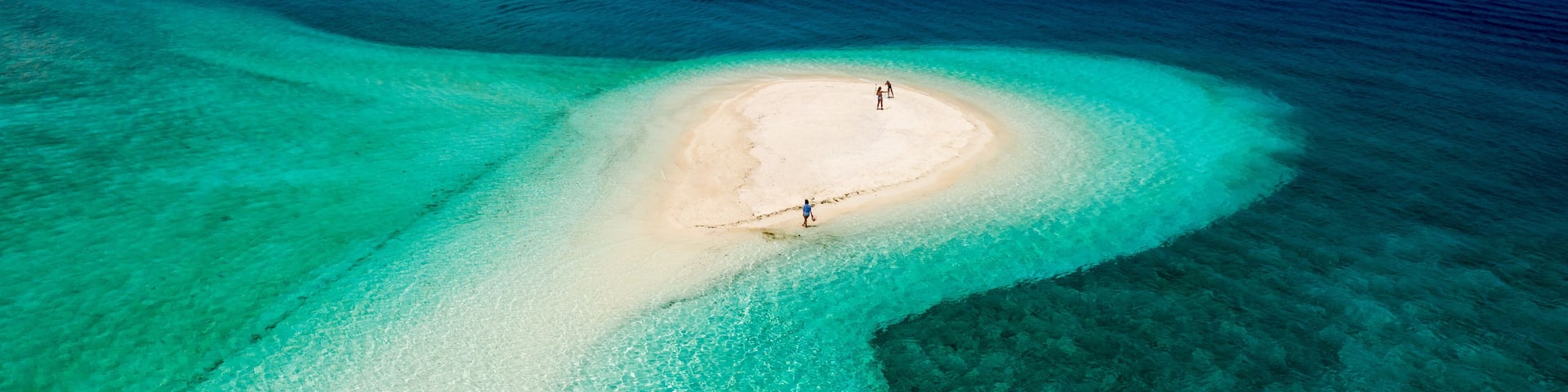 A clear aerial view of a turquoise lagoon with a sandy islet and two people standing on it.