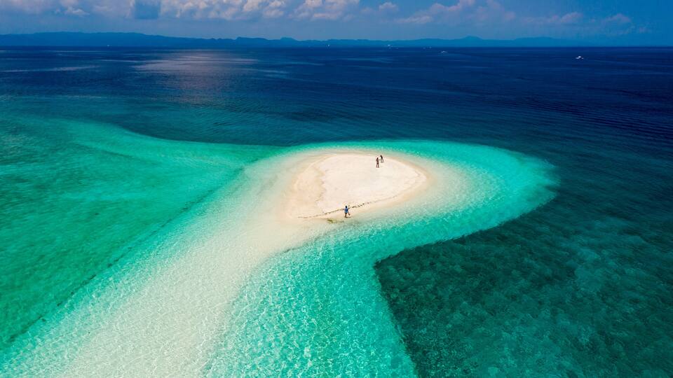 A clear aerial view of a turquoise lagoon with a sandy islet and two people standing on it.