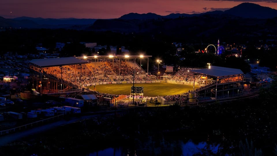 A stadium at dusk with floodlights, surrounded by mountains and a body of water.