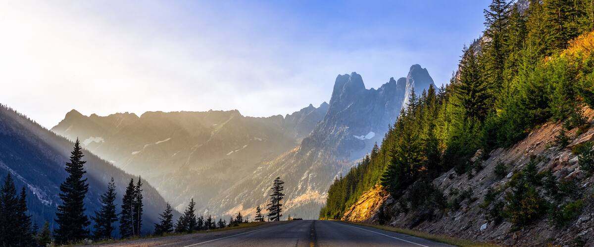 A mountain road flanked by evergreen trees and rugged peaks under a clear sky.