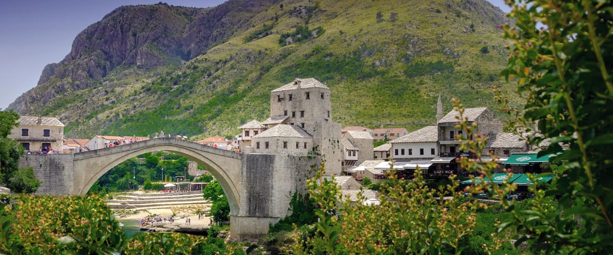 A stone bridge over a river, a town with traditional buildings, and a hill with a cross in the background.