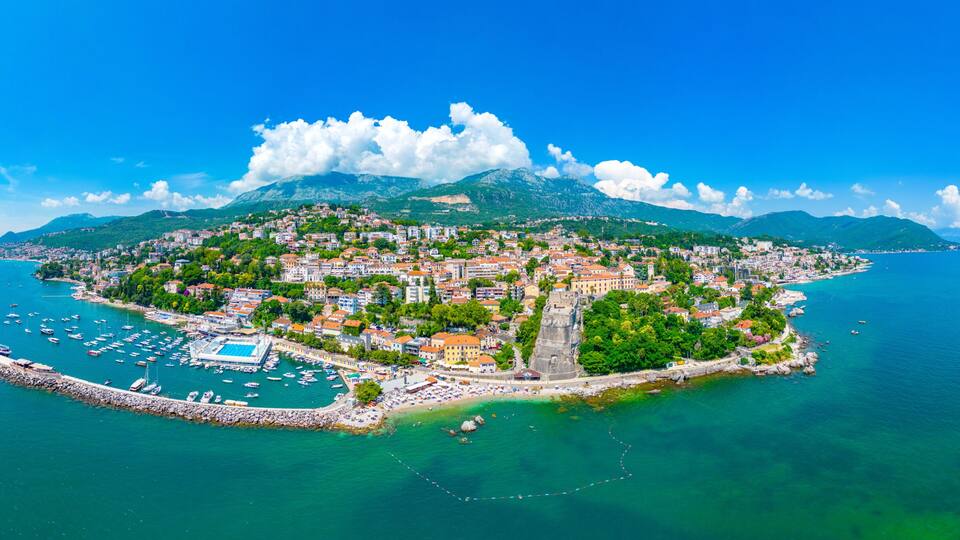 A coastal town with a marina, historic buildings, and a mountainous backdrop.