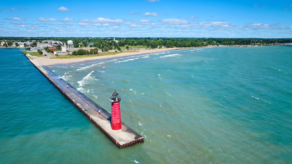 A lighthouse on a pier extending into a calm sea, with a beach and buildings in the background.