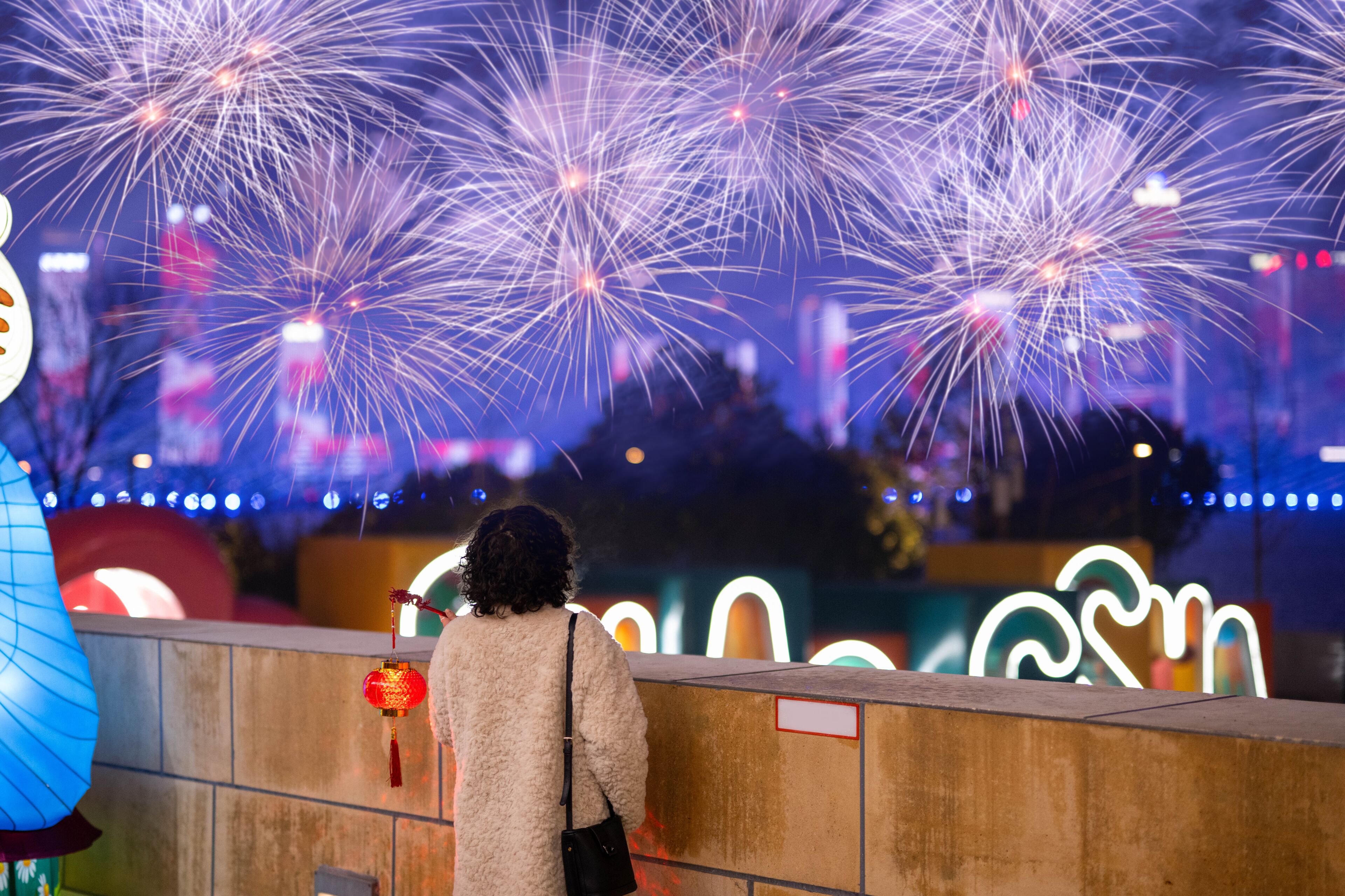 A person standing in front of a wall with a neon sign, watching fireworks in the background.