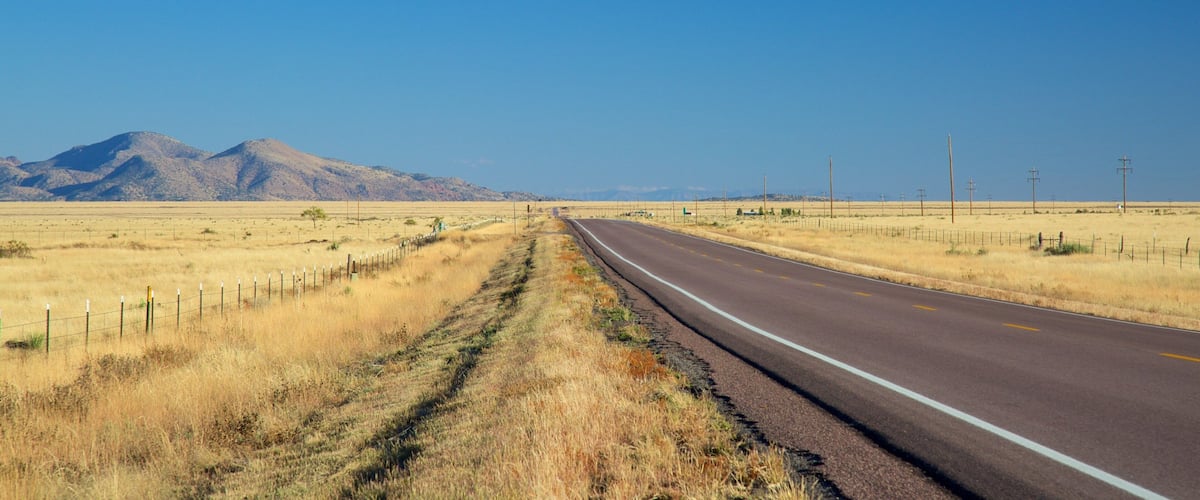 A straight road in a dry, grassy landscape with mountains in the distance and utility poles lining the road.