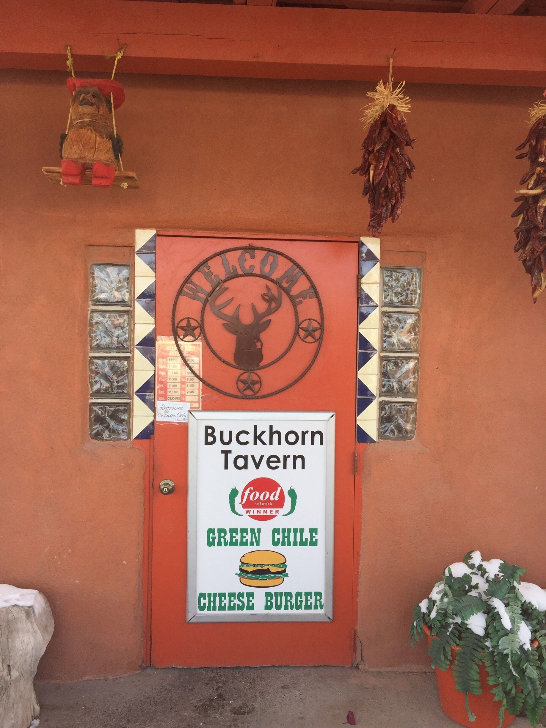 A red door with a 'Welcome' sign and a 'Buckhorn Tavern' sign advertising a green chile cheese burger.