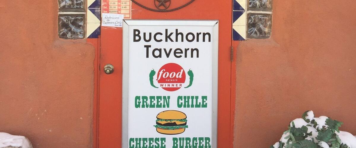 A red door with a 'Welcome' sign and a 'Buckhorn Tavern' sign advertising a green chile cheese burger.