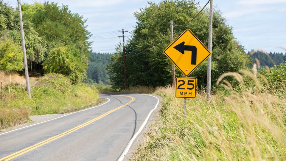 Long country highway with yellow lines down the centre and a caution sign indicating a left curve.