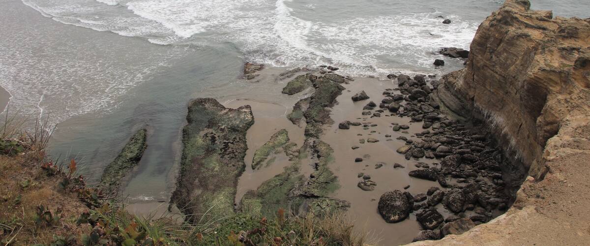 A coastal landscape with rocky cliffs, waves crashing onto the shore, and a distant ship on the horizon.