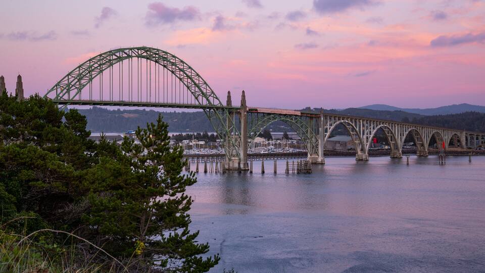 A steel arch bridge over a calm river with a backdrop of mountains and a sunset sky.
