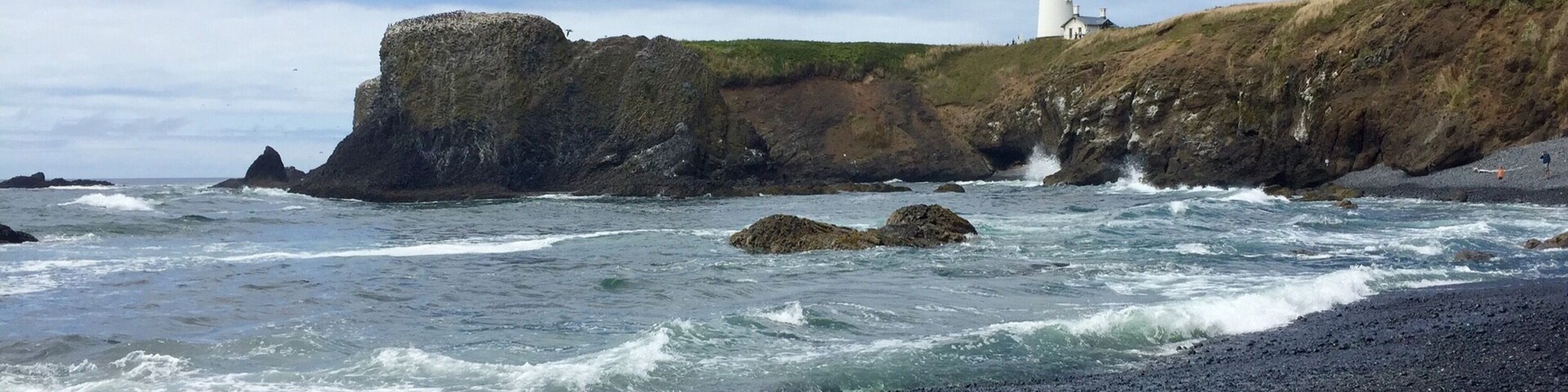 Costa rocosa con un faro en un acantilado, olas rompiendo contra la orilla y una playa de guijarros.