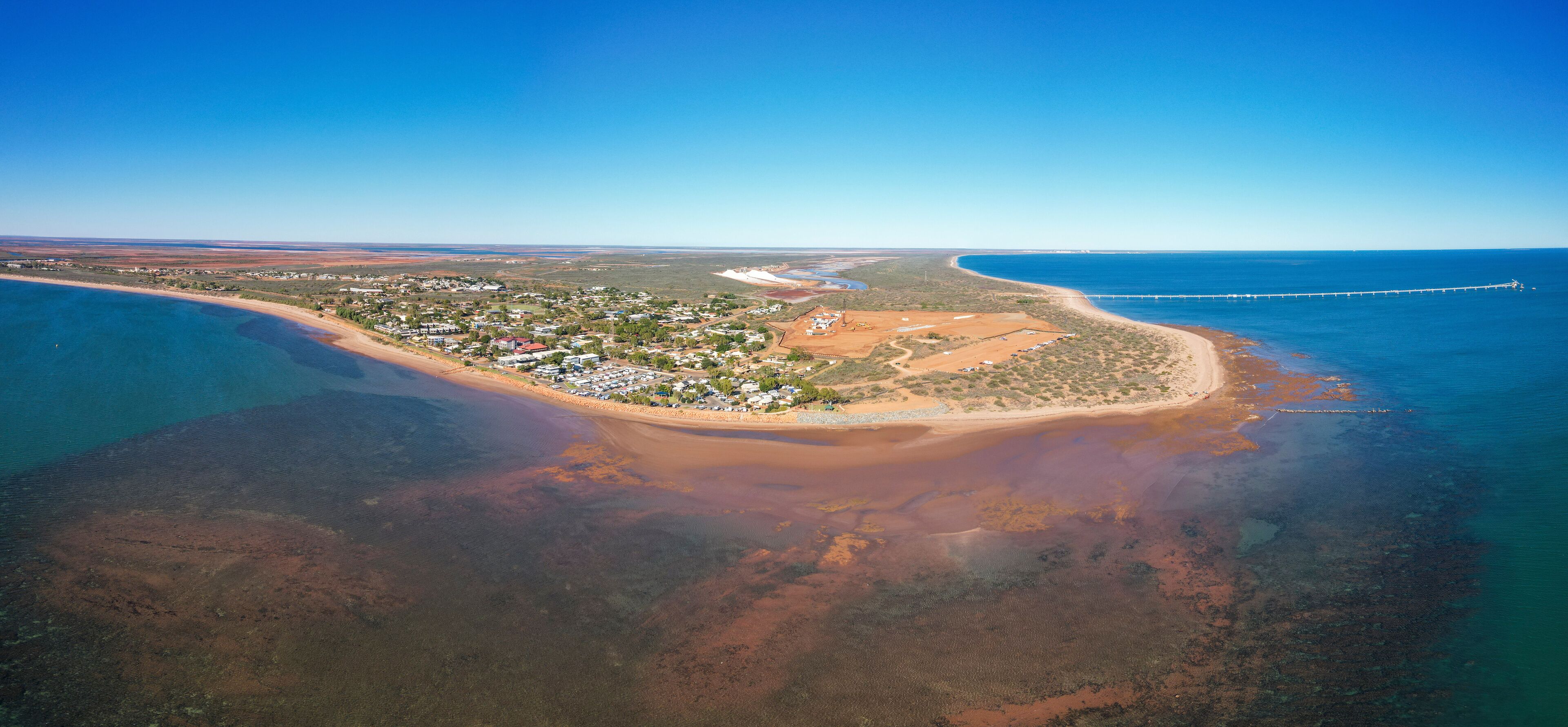 An aerial view of a coastal town with a beach, clear blue waters, and a long pier extending into the sea.