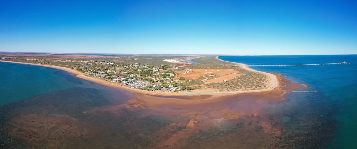 An aerial view of a coastal town with a beach, clear blue waters, and a long pier extending into the sea.