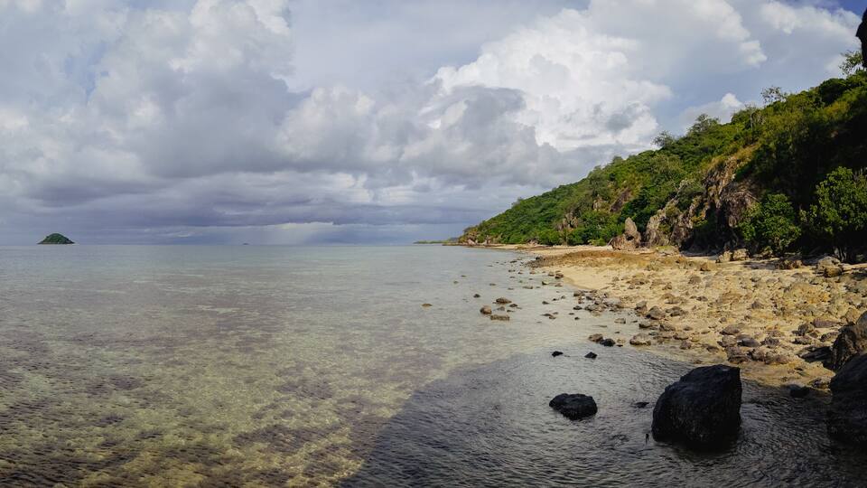 A beach with clear shallow waters, a distant island, and a rocky shoreline with vegetation.